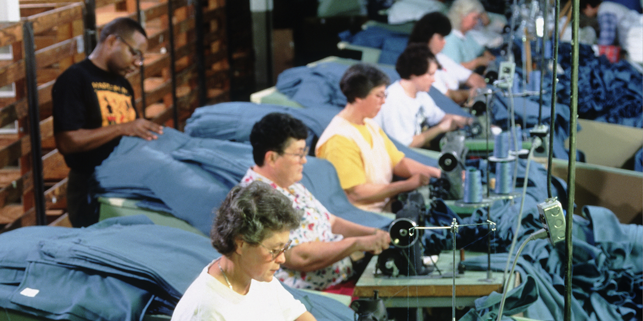 Photo of men and women sitting at sewing machines all in a row handling blue material Photo of men and women sitting at sewing machines all in a row handling blue material